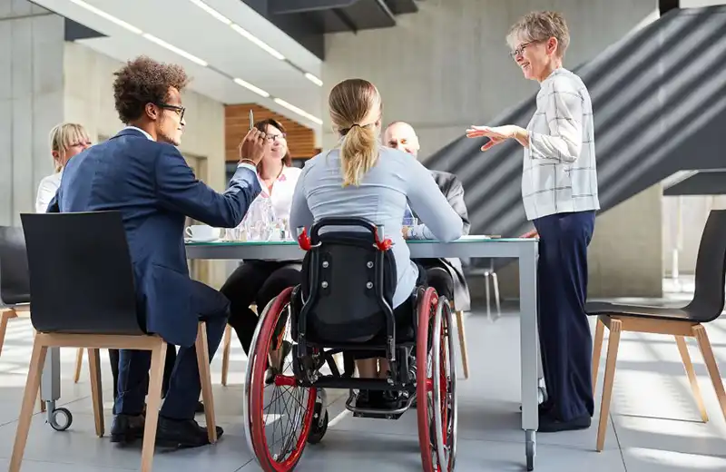 Group of diverse professionals having a meeting in a modern office, with one woman in a wheelchair participating actively and another woman standing and speaking.