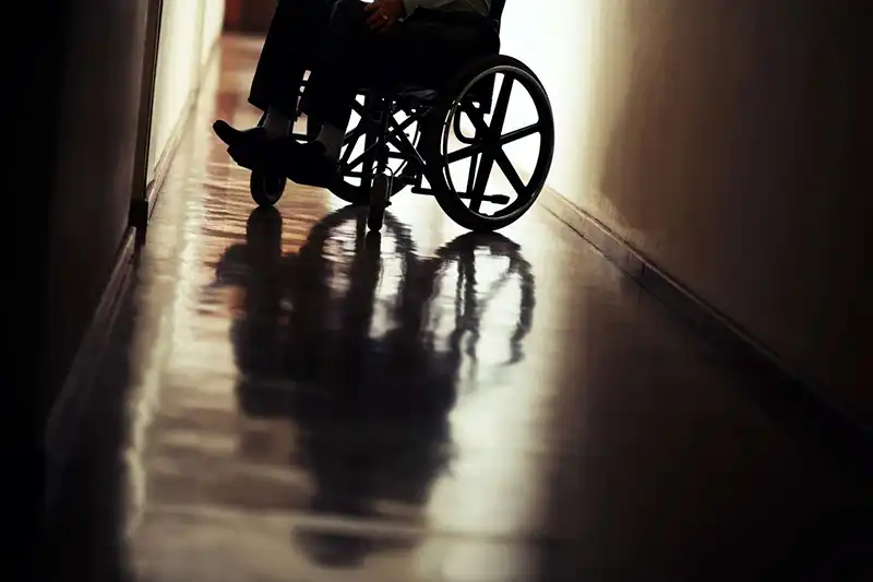 Silhouette of a person sitting in a wheelchair in a dimly lit hallway, with their reflection visible on the shiny floor.