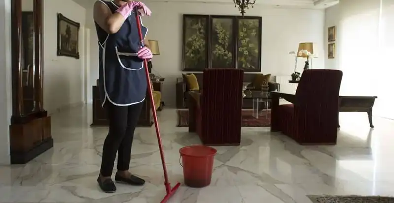 Person wearing pink gloves and a dark cleaning apron mopping the floor of a spacious, well-furnished living room with a red mop and a red bucket.