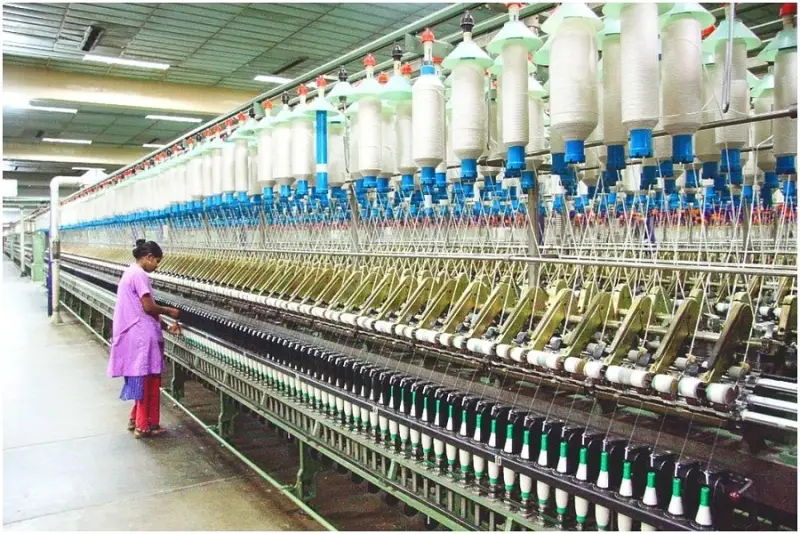Worker in a purple outfit operating textile machinery in a large factory with rows of thread spools.