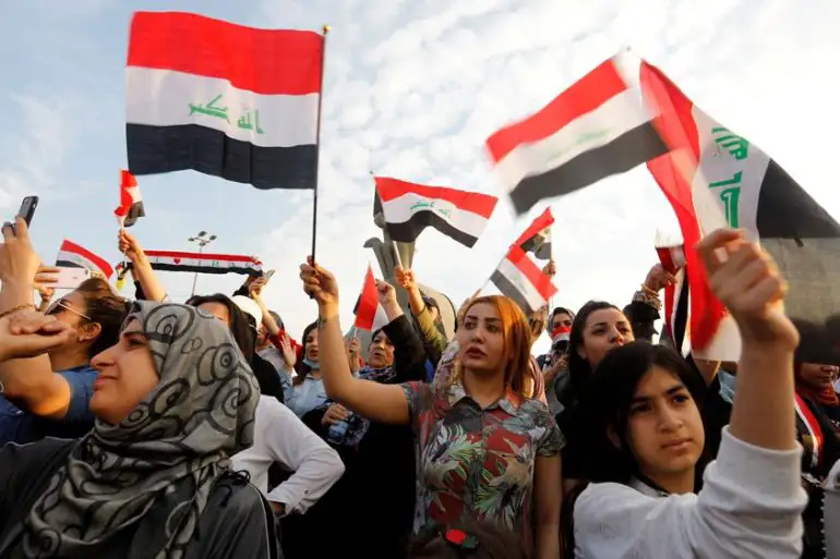 Crowd of people, mostly women, holding and waving Iraqi flags during a public gathering or protest.