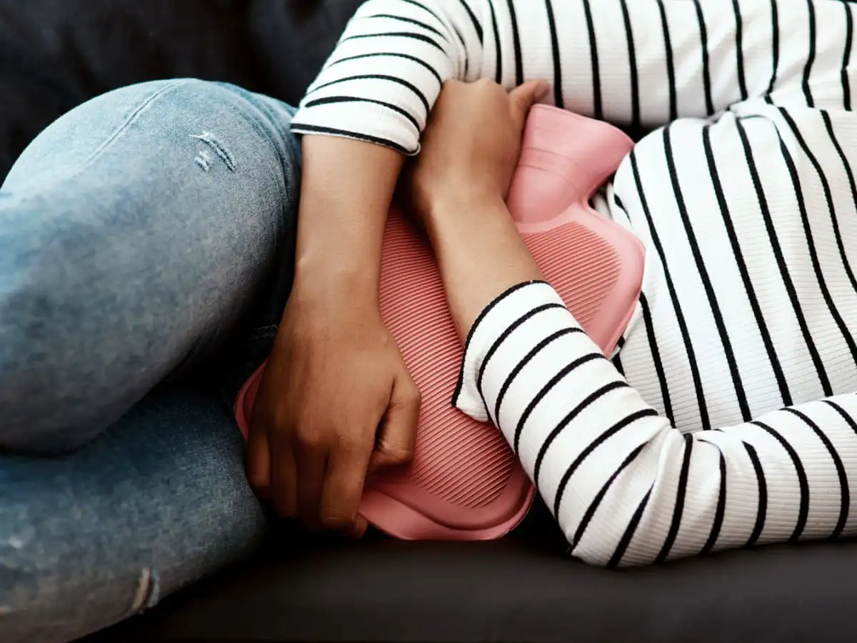 Person wearing a striped shirt and jeans holding a pink hot water bottle against their abdomen, possibly indicating pain or discomfort.