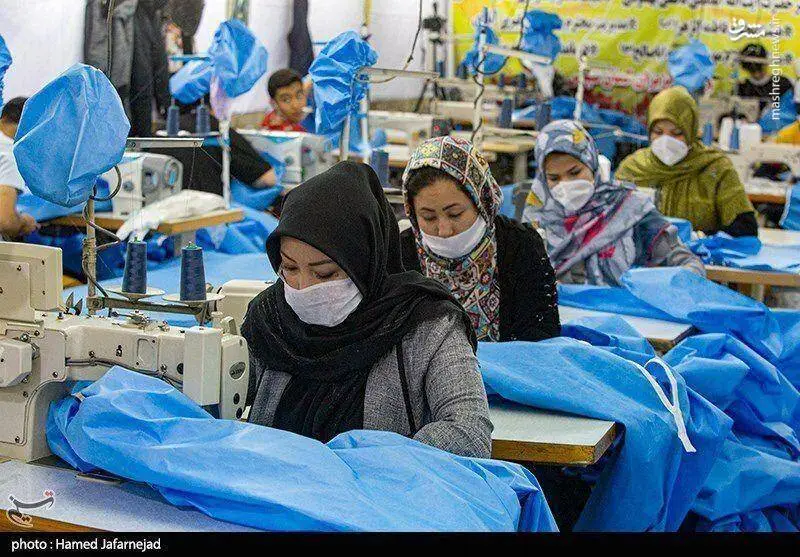 Women wearing headscarves and face masks working on sewing machines in a garment factory, stitching blue fabric.