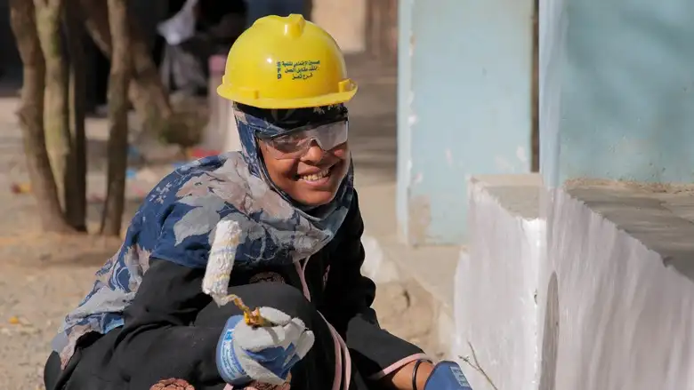 Woman wearing a yellow safety helmet and protective glasses smiling while painting a low wall with a roller brush.