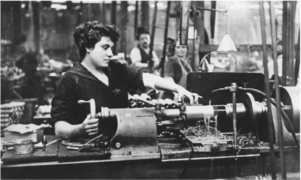 Black and white historic photo of a woman operating a machine in an industrial workshop with other workers blurred in the background.
