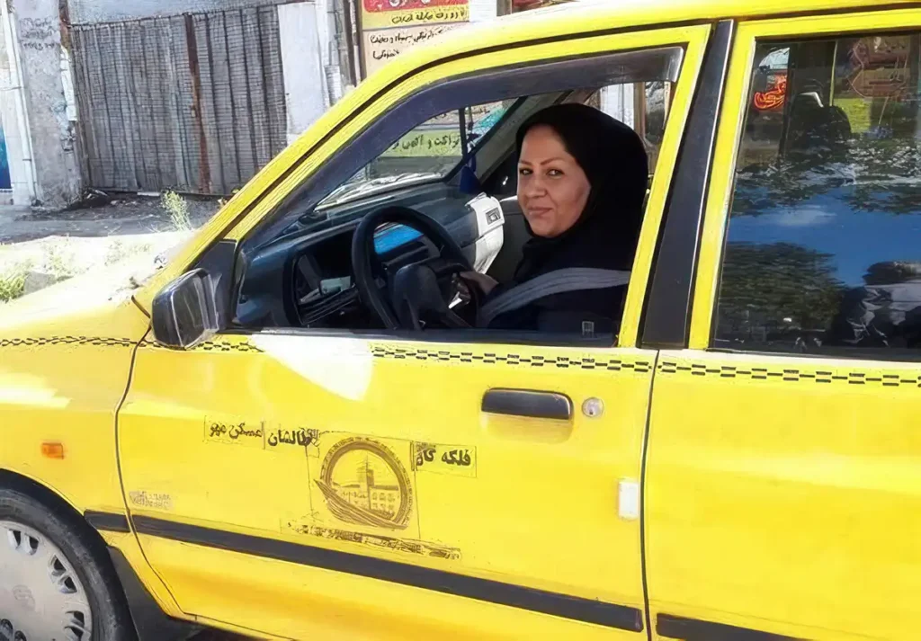 Woman wearing a headscarf and black clothes driving a yellow taxi, looking out the open window and smiling.
