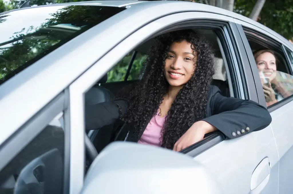 Smiling woman with long curly hair sitting in the driver’s seat of a car, with a happy female passenger in the back.