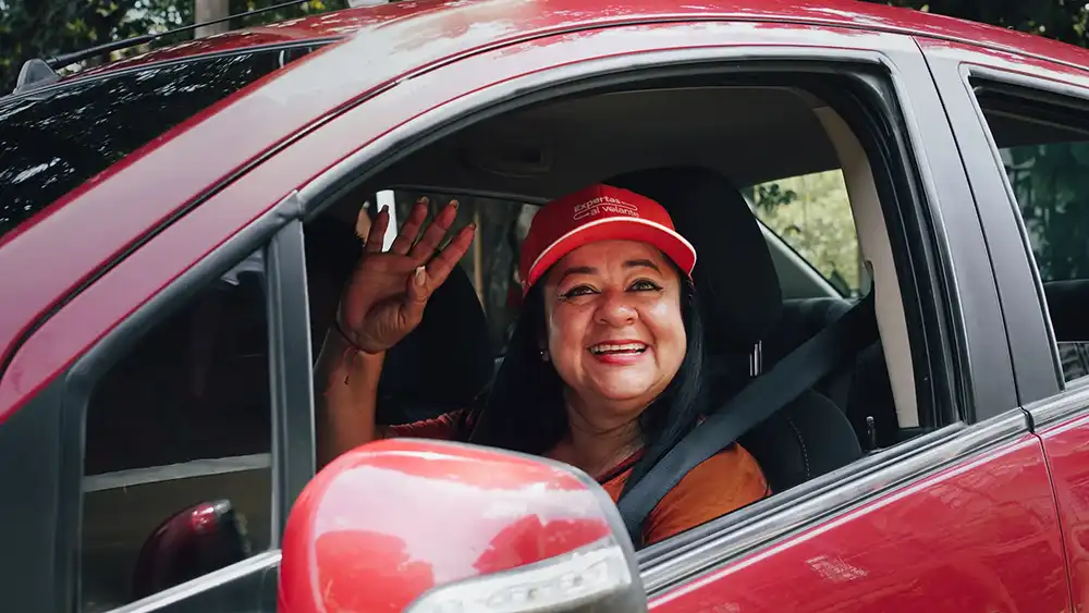 Woman in a red car smiling and waving from the driver’s seat, wearing a red cap and seatbelt.