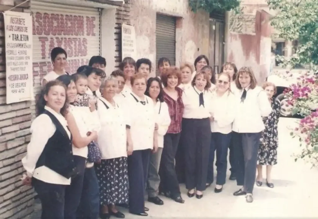 Group of women standing together for a group photo outside a building with a sign that reads "Asociacion de Taxis" in Spanish.  