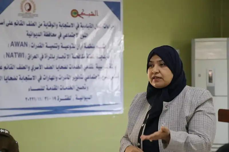 Woman wearing a headscarf and blazer standing and speaking in front of a poster with Arabic text.