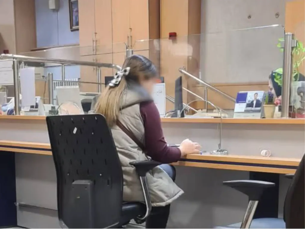 Person with long hair seated at a service counter in an office or bank, seen from behind interacting with staff across a glass partition.
