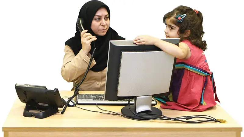 Woman wearing a headscarf at an office desk, speaking on a corded phone, with a young girl standing beside her and leaning on the desk.