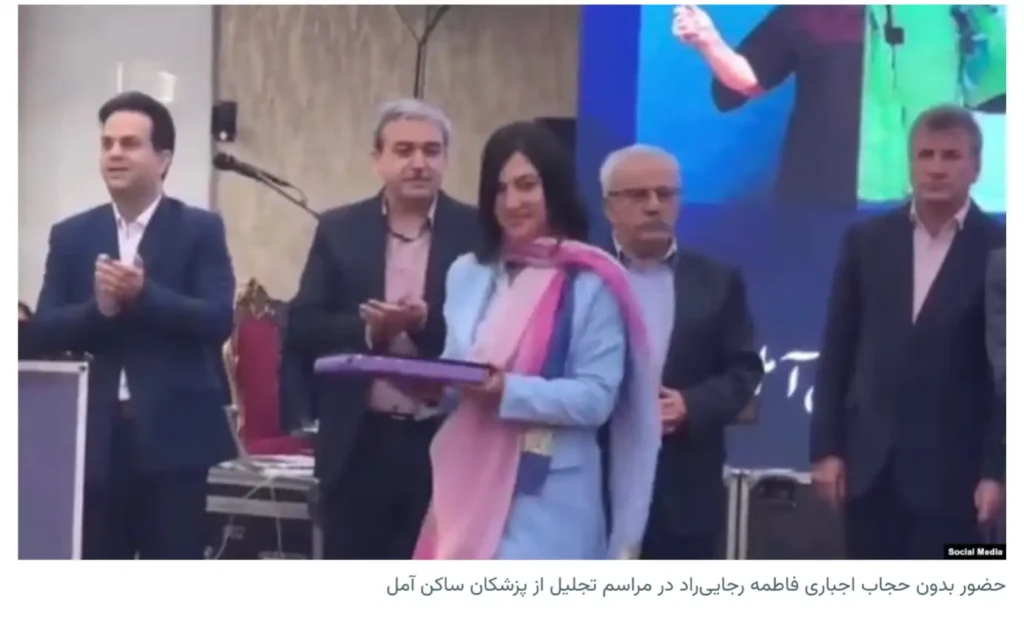 Woman without a head covering stands on a stage holding a wrapped plaque while several men in suits stand behind her and applaud; a Persian caption is visible below the photo.