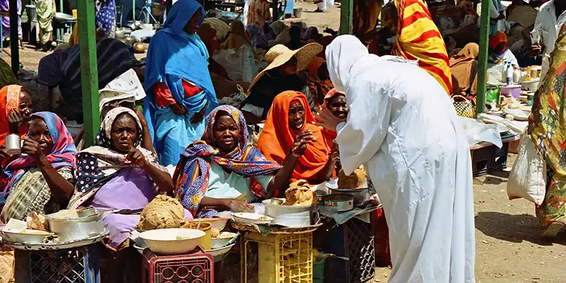 Busy outdoor market scene with several women sitting behind small makeshift stalls of bowls and pots of food, many wearing bright headscarves, while a standing vendor in white speaks with them.