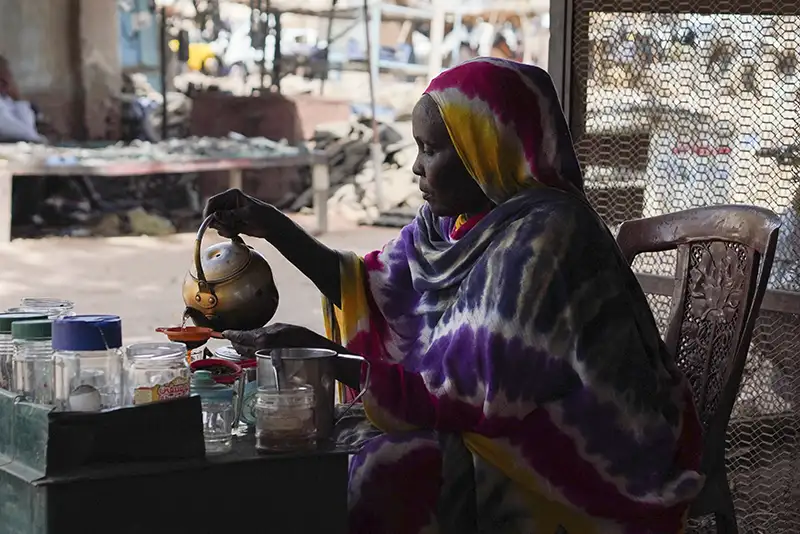 Woman in a tie-dye patterned headscarf seated at a table, pouring tea from a brass kettle into a small cup amid jars and utensils, light filtering through a metal screen behind her.