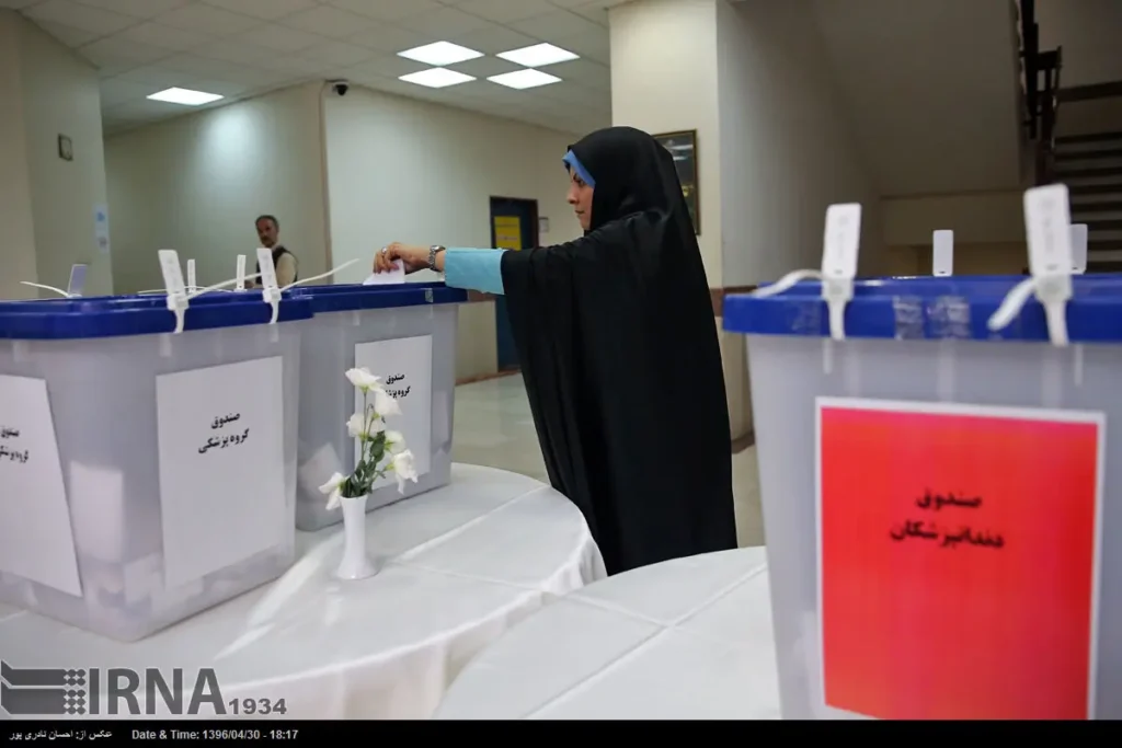 Woman wearing a hijab casting a ballot into a clear, sealed ballot box at a polling station, with multiple ballot boxes arranged on a table.  