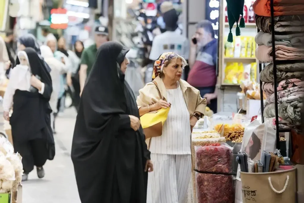 Busy market scene showing women browsing stalls and goods, including a woman in a black chador and an older woman wearing a headscarf examining items.
