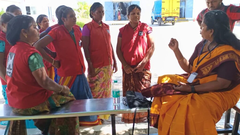 Group of women wearing red work vests and colorful skirts standing in a semi-circle listening as a woman in an orange‑yellow sari with a lanyard sits on a bench and gestures while speaking, with a blue truck and warehouse doors visible in the background.