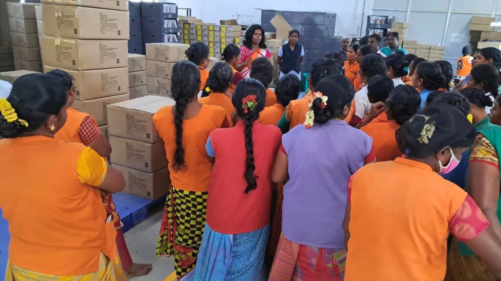 Photo of several women in red work vests standing outdoors while a woman in a yellow sari with a lanyard speaks to them, with a truck and building in the background