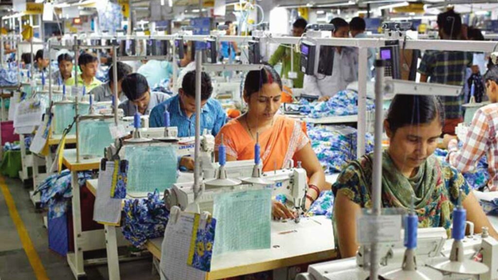 Photo of a garment factory assembly line showing rows of workers, mostly women, seated at sewing machines stitching fabric with stacks of boxes nearby.