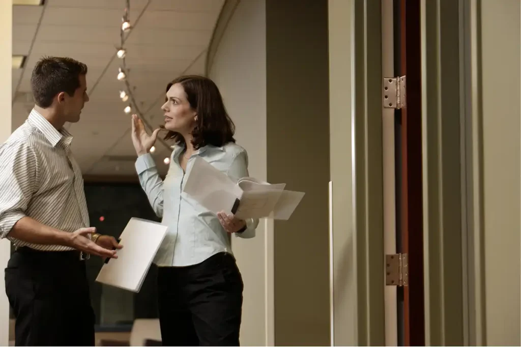 A man and a woman are standing in a doorway, engaged in an animated discussion or argument. The man, on the left, wears a striped short-sleeved shirt and dark pants, holding a white folder and gesturing with his hands. The woman, on the right, wears a light blue long-sleeved shirt and dark pants, holding papers in one hand and gesturing with the other raised. They are looking at each other. The background is an office hallway with blurred lights.