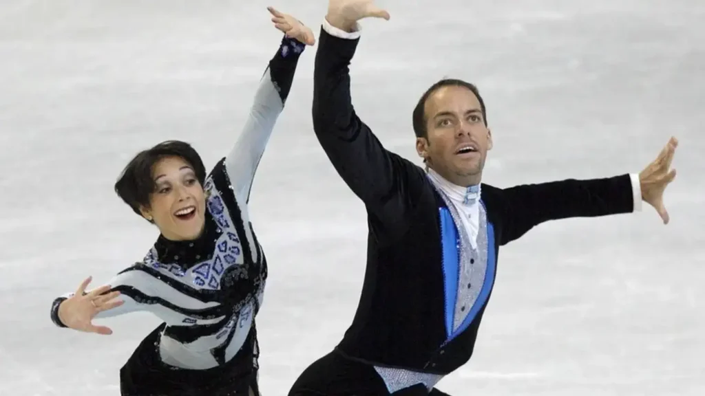 A male and female figure skater are performing on an ice rink. The female skater, on the left, has short dark hair and is wearing a black and white patterned costume, smiling with her arms outstretched. The male skater, on the right, has short dark hair and is wearing a dark blue and silver costume, with his arms raised and looking upwards.