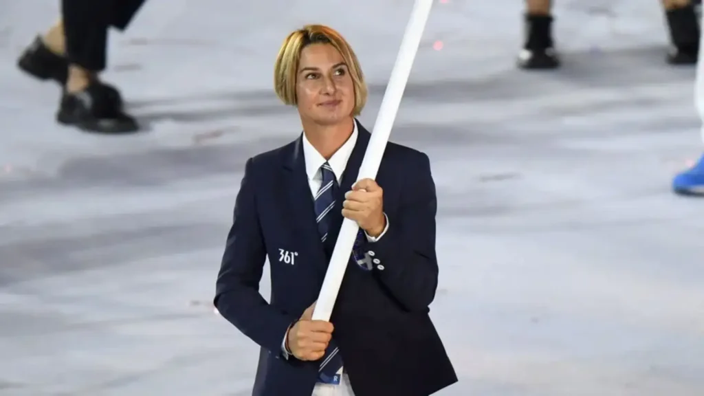 A person with short, light blonde hair is standing, wearing a dark blue suit jacket, a white shirt, and a blue and white striped tie. They are holding a white flag pole with both hands and looking forward with a slight smile. The background is blurred, suggesting they are at an event or ceremony.