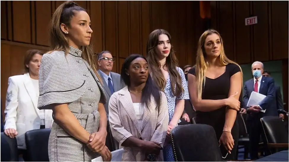 Four young women are standing side-by-side in what appears to be a formal setting, possibly a courtroom or hearing room. From left to right: the first woman has dark hair in a high ponytail and is wearing a patterned grey and white dress; the second woman, with long dark hair, is wearing a light-colored blazer and white top, looking downwards; the third woman has long brown hair and is wearing a blue and white patterned dress; and the fourth woman has long blonde hair and is wearing a black dress. There are other individuals, including men in suits, visible in the background.