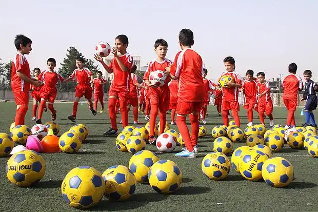 Numerous young boys, dressed in red soccer uniforms, are gathered on a green turf field under a bright sky. Many yellow and blue soccer balls, along with a few red and white ones, are scattered across the foreground and midground. The boys are actively engaged, some holding balls, others seemingly running or interacting with each other, suggesting a soccer practice or game.