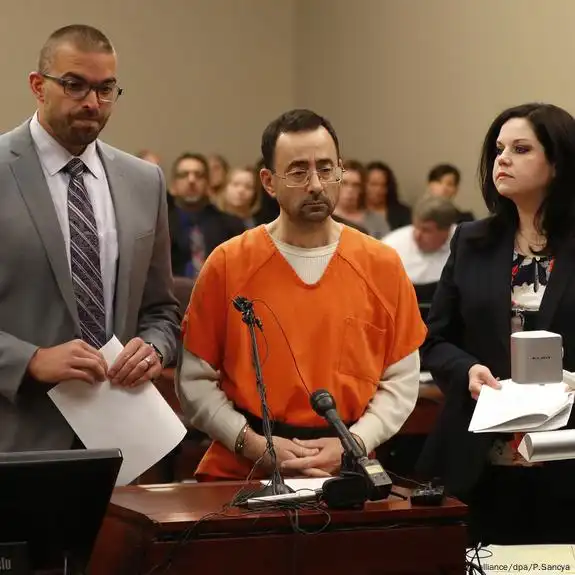 In a courtroom setting, a man in an orange jumpsuit is standing behind a wooden desk, looking forward with a somber expression. He has short dark hair and a beard. To his left, a man in a grey suit and striped tie is standing, holding papers. To his right, a woman in a dark blazer and patterned top is standing, also holding papers. A microphone is on the desk in front of the man in the jumpsuit.