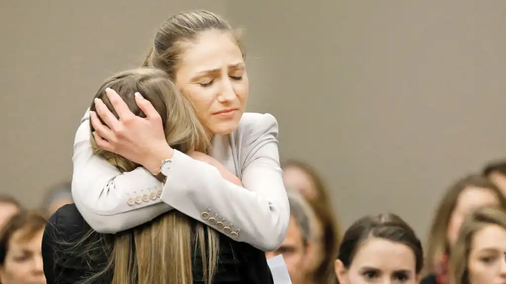 Two women are embracing each other tightly. The woman on the right has blonde hair tied back and is wearing a light-colored blazer, with her eyes closed in what appears to be distress or sadness. Her arms are wrapped around the other woman, whose back is to the viewer, showing long blonde hair. The background is blurred, indicating other people are present.