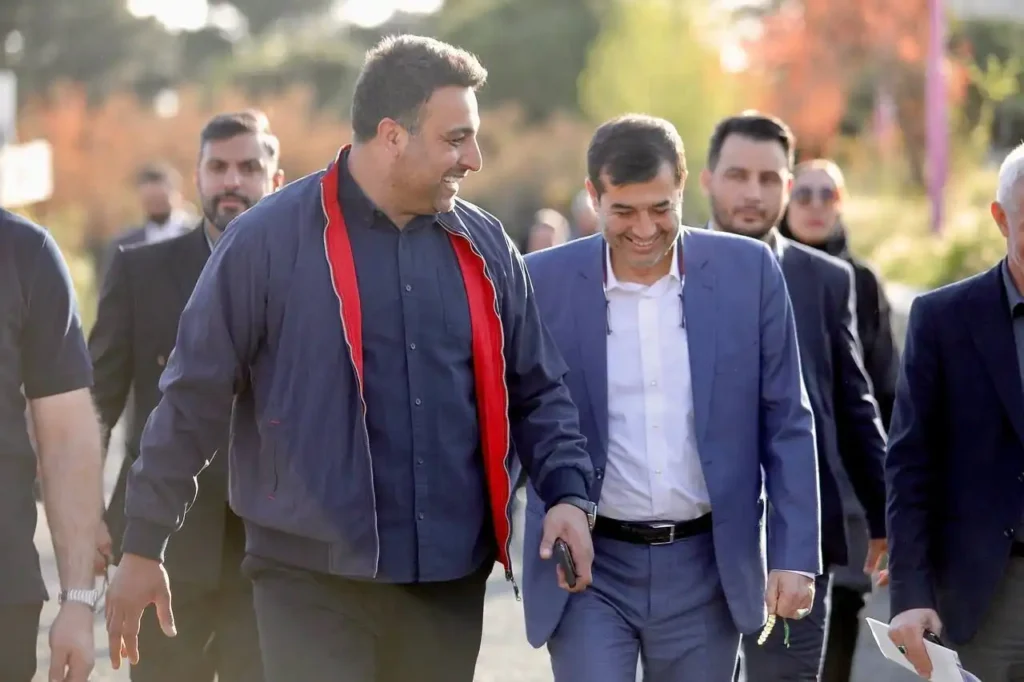 Two men are walking side-by-side and smiling, engaged in conversation. The man on the left is wearing a dark blue jacket with red lining and a dark shirt. The man on the right is wearing a light blue suit and a white shirt, holding what appears to be a phone in his left hand. Other blurred figures are visible in the background, suggesting an outdoor public setting with trees and sunlight.
