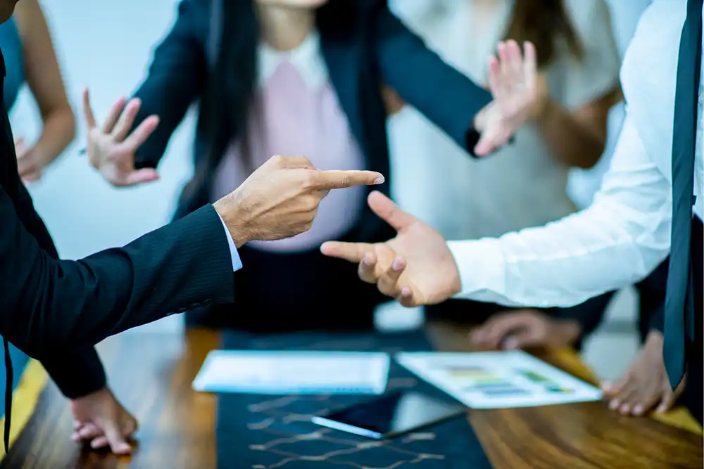 Close-up of two people’s hands pointing at each other over a table during a heated discussion, with blurred colleagues in the background making stopping gestures.