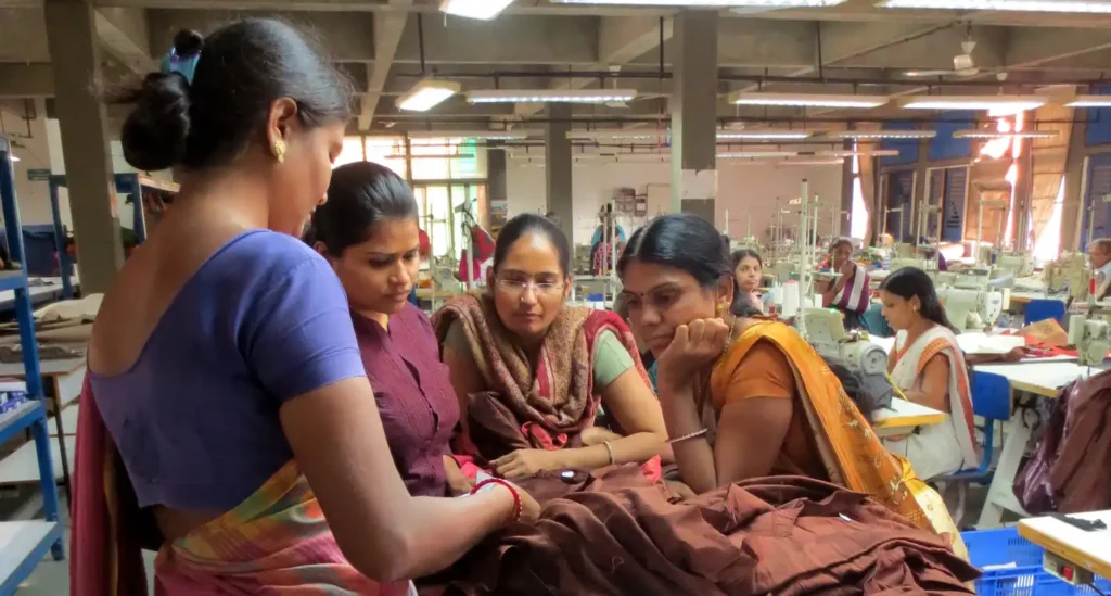 Four women are gathered around a table in what appears to be a garment factory or workshop, with sewing machines visible in the background. The woman on the far left, seen from behind, is wearing a blue top and a colorful sari. The other three women are looking down at a large piece of brown fabric on the table, seemingly inspecting or working on it. They are all wearing traditional Indian clothing.