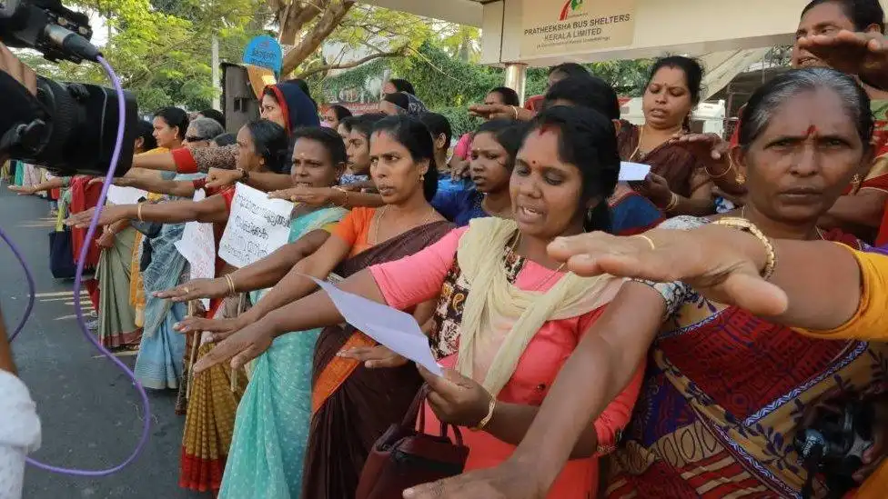 A large group of women are standing outdoors, many with their right arms extended forward at shoulder height, participating in a protest or demonstration. The woman in the center foreground is holding a piece of paper and appears to be speaking or chanting. Many are wearing colorful saris. A sign for "PRATHEEKSHA BUS SHELTERS, KERALA LIMITED" is visible in the background.