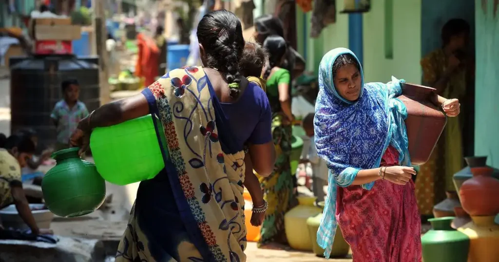 Two women are walking on an unpaved path, both carrying large water containers. The woman in the foreground, seen from behind, is carrying a bright green pot and a green bucket on her left side, and has a child on her back. She is wearing a blue and patterned sari. The woman in the middle ground, facing right, is wearing a blue patterned headscarf and a pink patterned dress, carrying a brown pot on her right hip. Other blurred figures and pots are in the background, suggesting a village or rural setting.