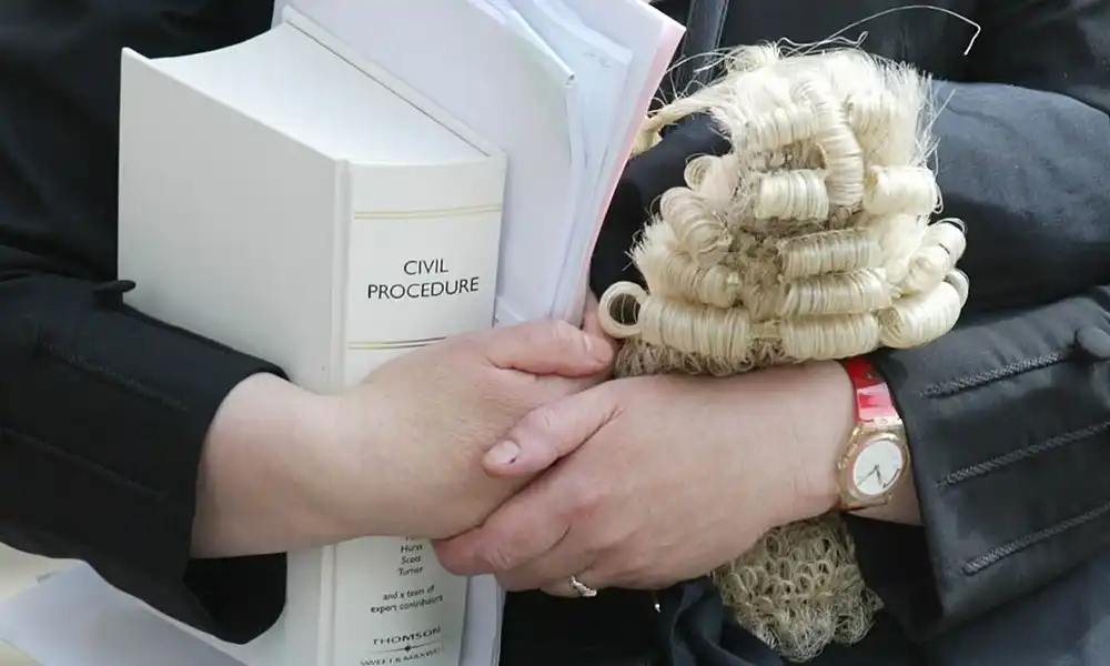 A close-up of a person's hands, wearing a black robe, holding a white legal textbook titled "CIVIL PROCEDURE" and a traditional blonde barrister's wig. The person is also wearing a watch with a red strap.