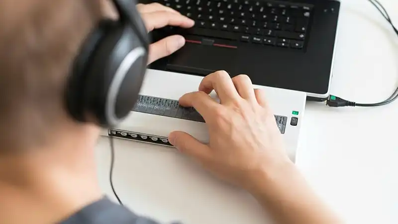 A close-up, over-the-shoulder view shows a person wearing black headphones, with their hands positioned over a white braille display connected to a black laptop. The person's fingers are touching the braille cells, and they appear to be reading or inputting information using the device.