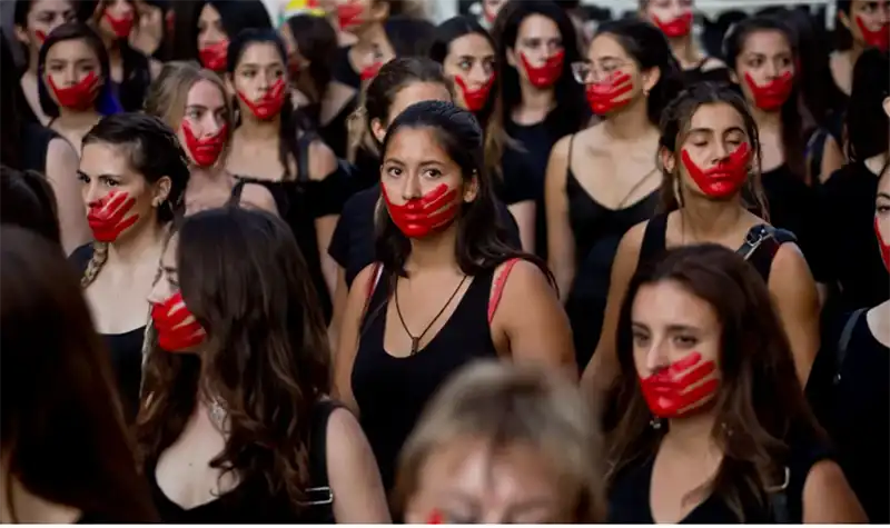 A large group of women are gathered, many of whom have red paint resembling a handprint covering their mouths. They are mostly wearing dark clothing. The woman in the center foreground is looking directly at the camera with a serious expression, and a red handprint is visible on her mouth.