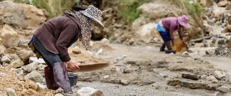 Two women are seen engaged in what appears to be informal mining or sifting activities in a muddy, rocky landscape. The woman in the foreground is bent over, wearing a wide-brimmed patterned hat, long-sleeved brown top, dark pants, and tall boots, sifting material in a shallow pan. In the background, another woman in similar attire is also bent over, working in the mud.
