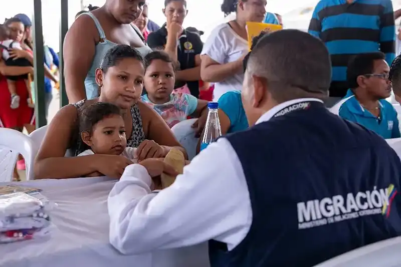 A woman with dark hair, holding a small child, is sitting at a white table facing a man in a white shirt and dark blue vest with "MIGRACIÓN" written on the back. The man is speaking to her across the table. Another child is sitting next to the woman, and other people are visible in the blurred background, suggesting a processing or assistance center.