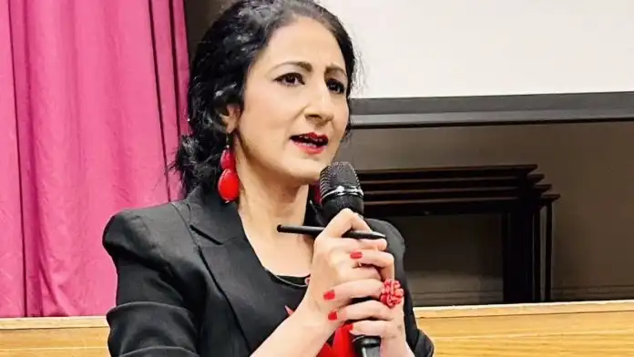 A woman with long dark hair, wearing red teardrop earrings and a black blazer, is speaking into a microphone with red-painted fingernails holding it. Her lips are slightly parted, and she has a focused expression. A pink curtain is visible in the background.