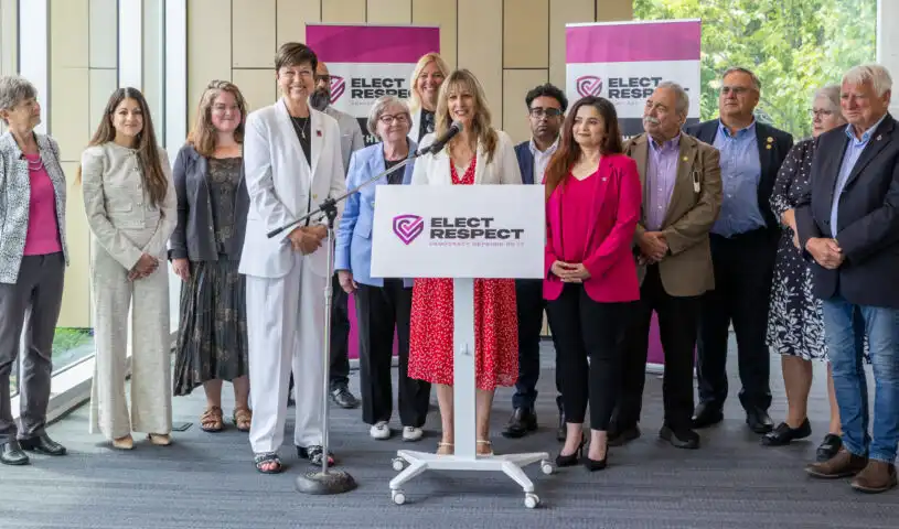 A diverse group of about fifteen people, including many women, are standing behind a white podium with the logo "ELECT RESPECT DEMOCRACY DEMANDS IT". A woman in a red and white polka-dot dress is speaking at the podium. Other individuals are standing to her left and right, some smiling. Two pink banners with the same logo are in the background.