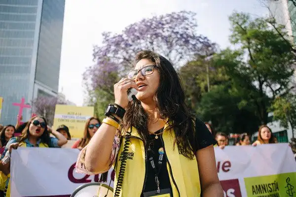 A woman speaking into a megaphone at a protest, wearing a yellow Amnesty International vest, with other demonstrators and banners behind her in a city street.
