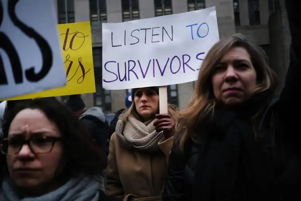 A crowd of protestors holding signs, including one that reads “LISTEN TO SURVIVORS,” during a public demonstration in an urban setting.
