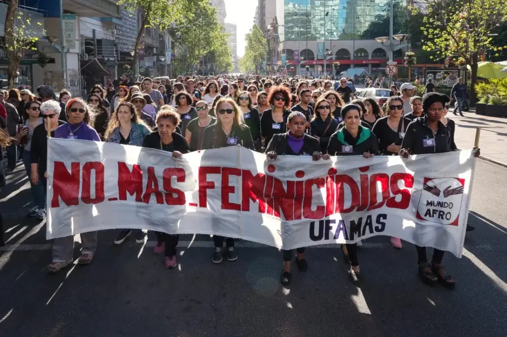 A large group of women marching in a street protest, carrying a banner that reads “NO MÁS FEMINICIDIOS” and “UFAMAS,” with “Mundo Afro” visible on the right.
