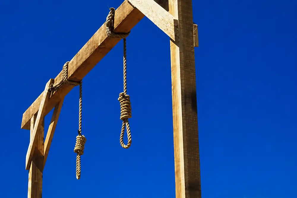 A wooden gallows structure photographed from below against a clear bright blue sky, with two rope nooses hanging from the horizontal beam.