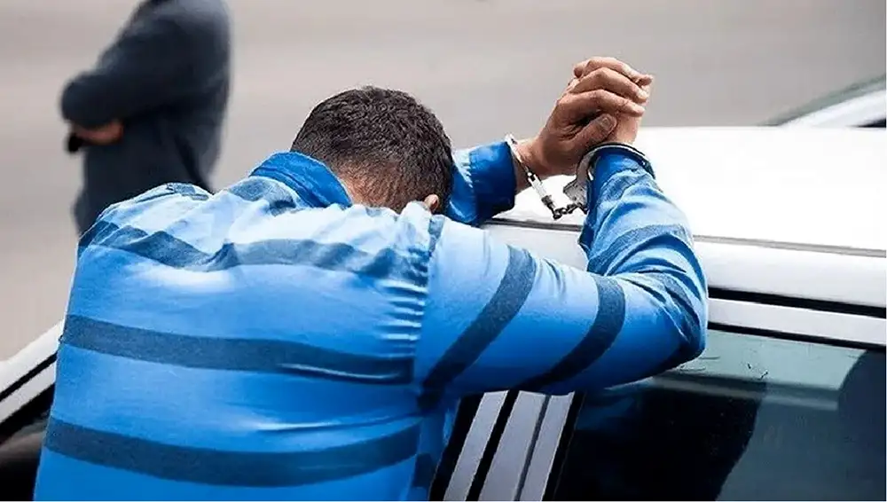 A man in a blue striped shirt, hands handcuffed behind his back, leaning face-down against the roof of a car. A second person stands blurred in the background.