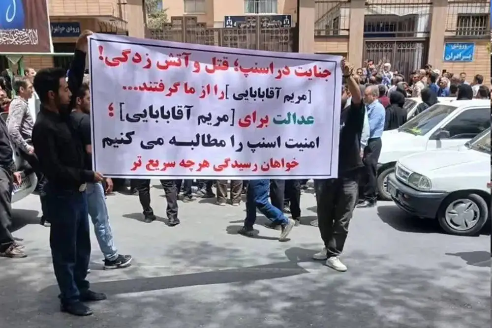 A crowd of protesters on a street holding up a large white banner with red and black Persian text referencing Maryam Aghababaei, demanding justice and safety. Buildings and parked cars are visible in the background.